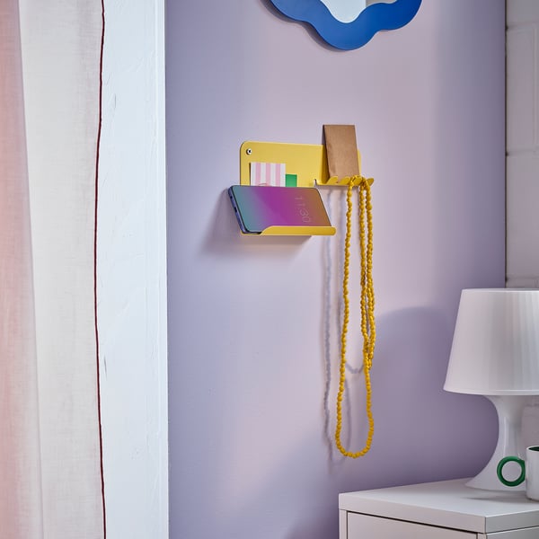 Small yellow steel wall organiser with shelf, holding phone, keys, and beads, mounted on purple wall.