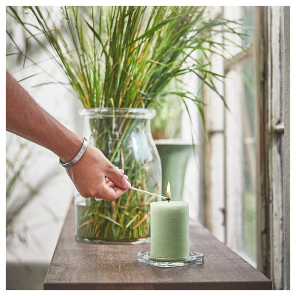 A person lights a green HEDERSAM candle on a wooden shelf, next to a large glass vase with long green grasses.