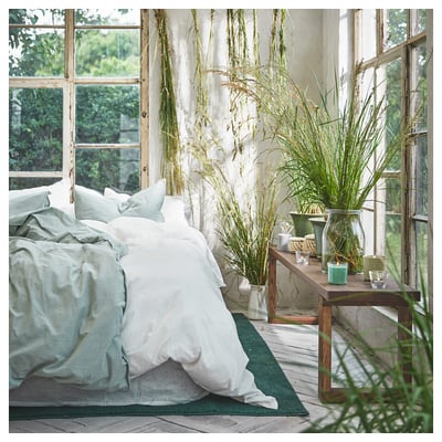 Bright, sunlit bedroom with white bedding, tall green plants in vases, and large windows offering natural views.
