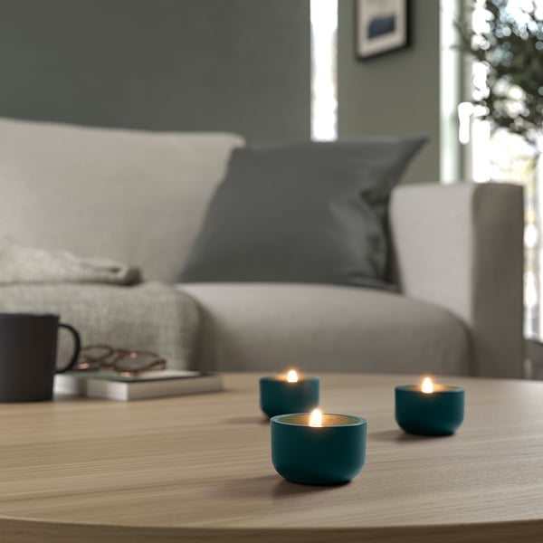 Three lit tealights on wooden table, white couch with black pillows, cosy natural light.