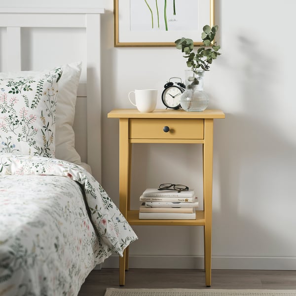 Small yellow HEMNES bedside table with drawers and shelves holds items neatly.