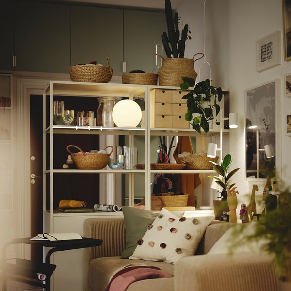 Cosy living room with a beige HÖKRUBBA round woven basket on a shelf surrounded by plants, decorative items, and a green throw pillow on a couch.