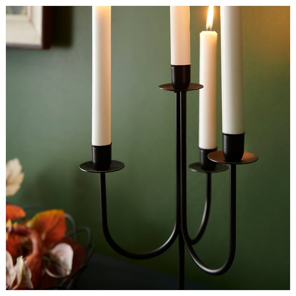 Black metal candelabra with three white candles. One candle is lit, casting soft light. Placed on green table with flowers.