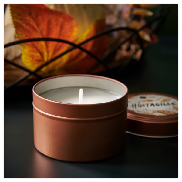 Round tin scented candle, lit, on dark surface. Label reads HÖSTAGILLE. Warm, inviting ambiance with orange and yellow leaves in background.