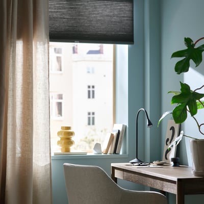 Home office with blue walls, grey blinds, wooden desk, lamp, chair, plant, and yellow vase.