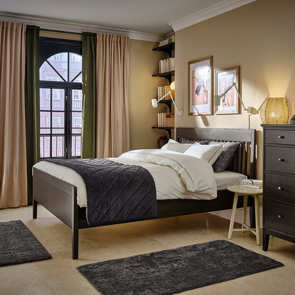 Bedroom with dark wood IDANÄS bed, white linens, black and grey rugs, black dresser, round lamp, picture frames, large window with brown curtains.