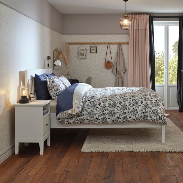 Cosy bedroom with white bed, dark floral bedding, wooden floors, rug, and pink curtains.