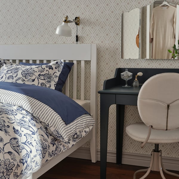 Elegant white wooden bed with blue floral bedding, wall-mounted lamp, black vanity desk with beige chair, patterned wallpaper, mirror, and clothes.