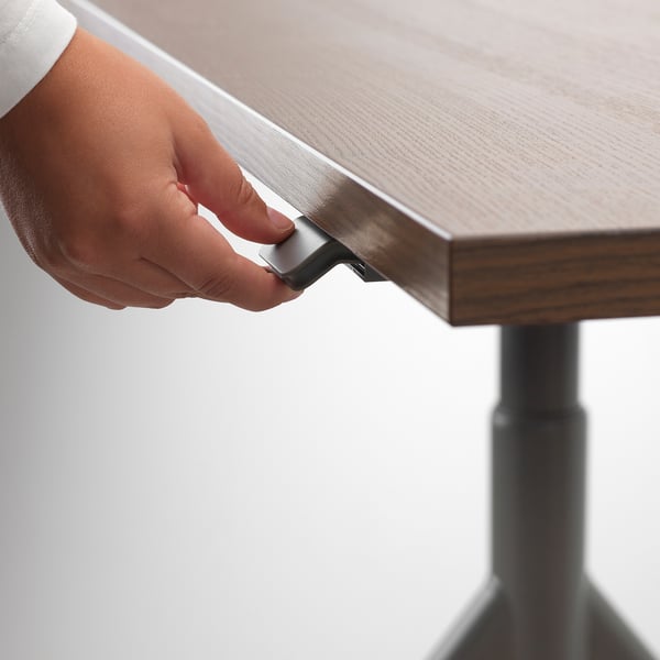 Person adjusting height of IDÅSEN desk using a handle. Desk has adjustable height and cable management.