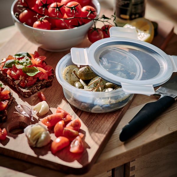 Wooden cutting board with knife, holding a plastic food container with leftovers, and a bowl of cherry tomatoes.