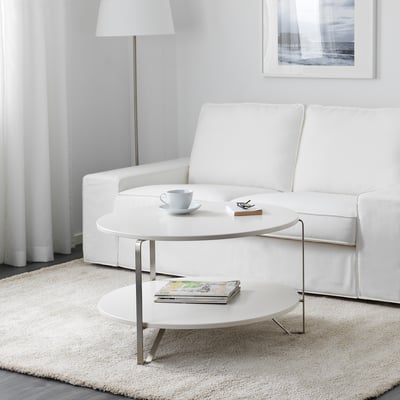 Modern living room with white furniture. Round coffee table has a lower shelf for magazines and an organised top with a cup and remote.