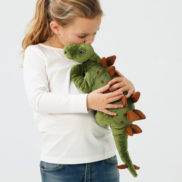 A girl holds a green plush dinosaur toy. Shes hugging it close, showing its comforting and cuddly nature. The toy has brown spots and spikes.