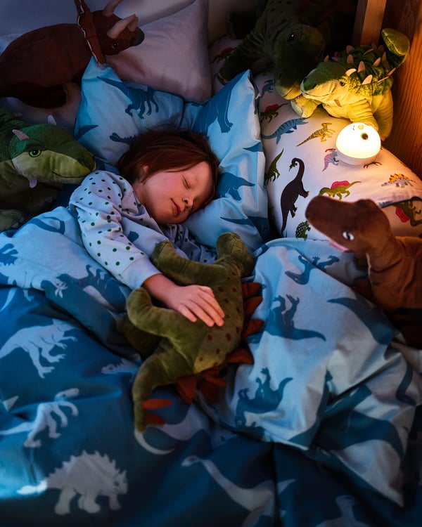 A child sleeps on a dinosaur-themed bed with green and brown stuffed dinosaurs, blue sheets, and a nightlight.
