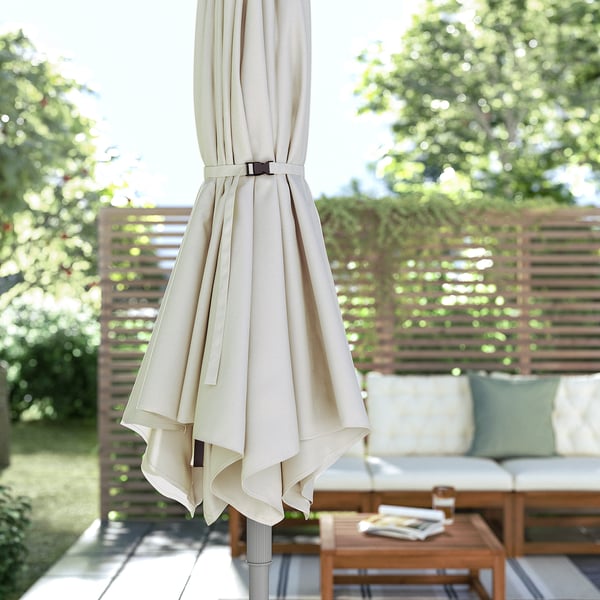 Closed beige outdoor umbrella, JOGGESÖ, leaning on patio furniture, surrounded by greenery.