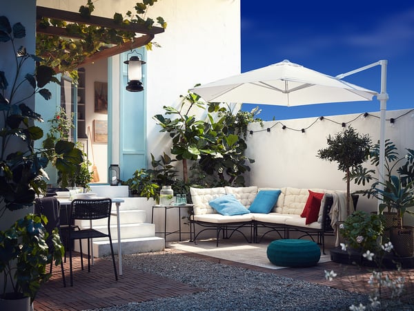 Outdoor patio with JUTHOLMEN white sofa, blue pillows, brick floor, plants, and white umbrella.