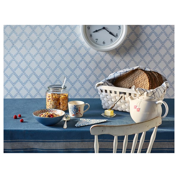 A cosy breakfast setup with a bowl of cereal, a white teapot, sliced bread, and a chair against a blue and white patterned wall.