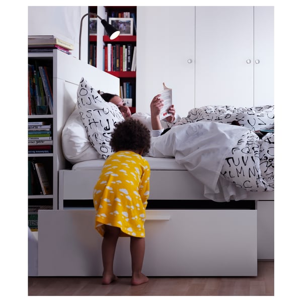 Person reading in bed, child in yellow dress, bookshelf headboard, adjustable lamp.