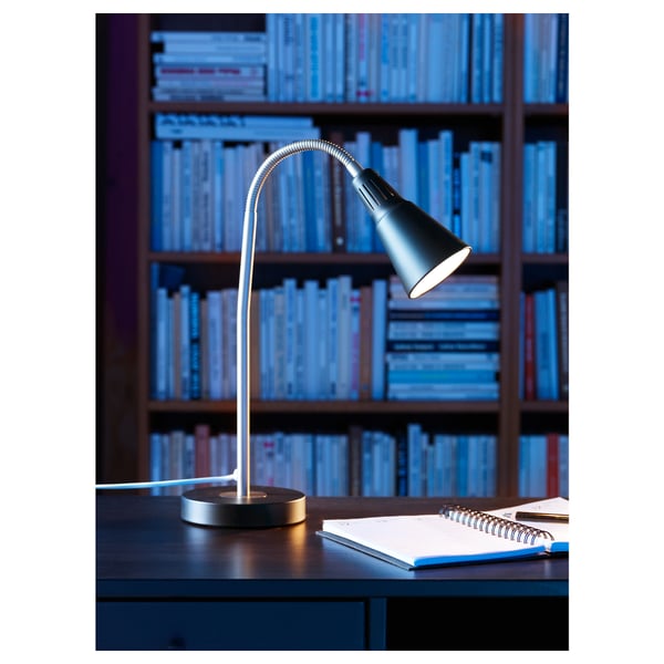 A black adjustable desk lamp with a flexible neck is lit on a table in front of shelves filled with books. A notebook is also on the table.