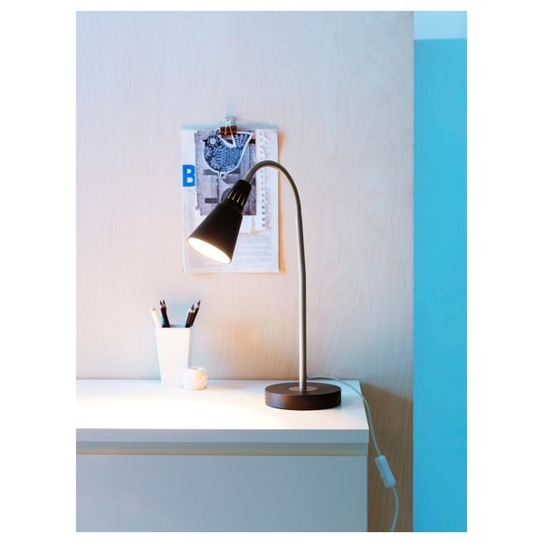A black KVART desk lamp sits on a white table, emitting light downwards. Pencils in a cup and a clipboard are nearby.