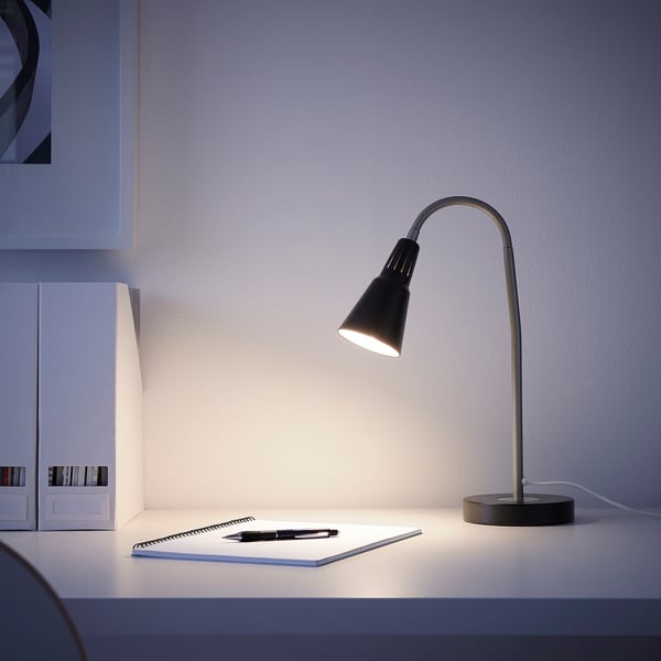 Small black KVART desk lamp illuminates a white table with a notebook and pen, casting light beside a white box.