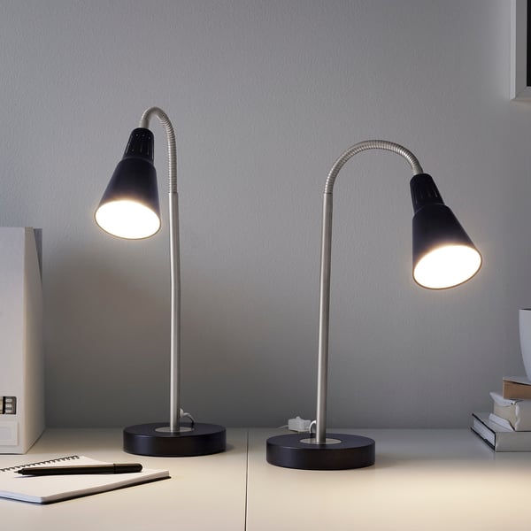 Two black KVART lamps on a desk. Flexible neck for directional light. Smaller than nearby books and notebooks. Grey wall background.