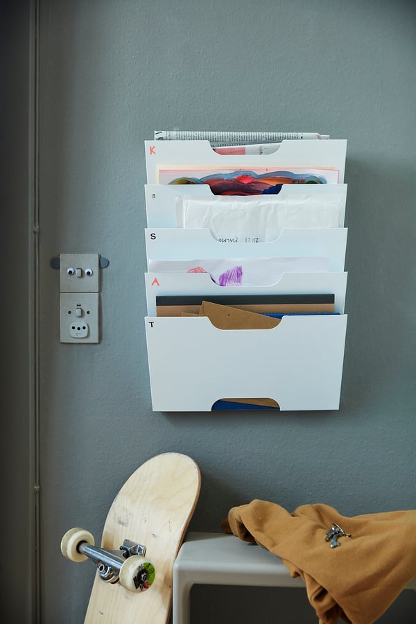 White KVISSLE wall file organiser with coloured folders on grey wall near wooden skateboard.
