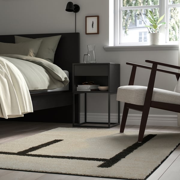 Modern bedroom with cream bed, black bedside table, armchair, and LÄNSVÄG off-white rug.