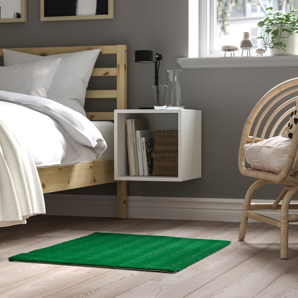 Modern bedroom with LANDSVÄG green rug, white bed, wicker chair, and wall storage.