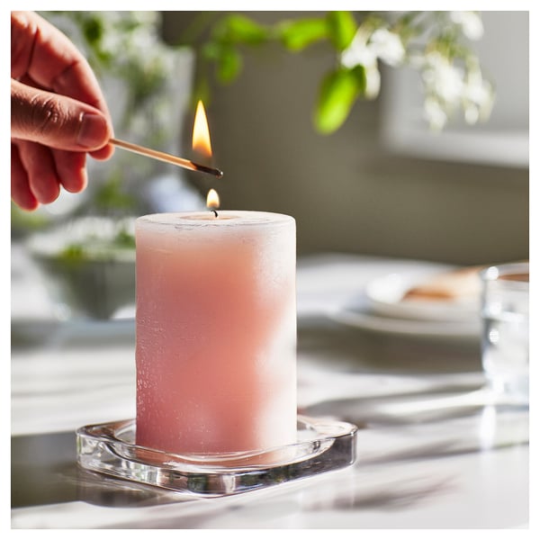 A hand lights a white LUGNARE scented candle on a glass holder, set on a table with blurred green plants in the background.