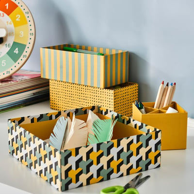 Desk with 4 LYSMASK boxes: yellow, green, black. Feathers, scissors, coloured pencils inside. Stacked, recycled.