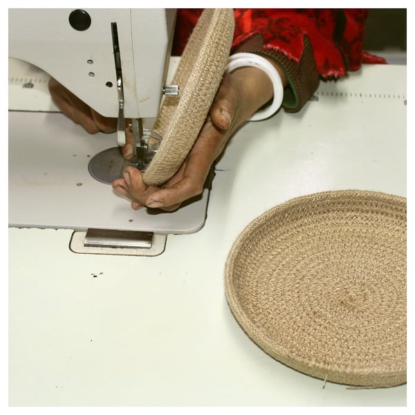 Person operating sewing machine, crafting MÄVINN basket. Beige jute and cotton; round, hand-woven.