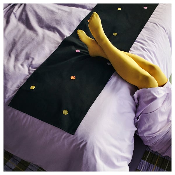 A black cotton bed runner with colourful crocheted dots, featuring yellow legs protruding from under it.