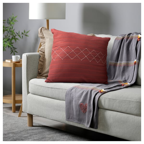 A cosy, red, rectangular MÄVINN cushion on a grey couch, with a geometric hand-embroidered pattern, next to a lamp and a grey throw with tassels.