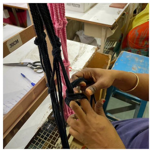 Hands craft black cotton macramé plant hanger, showcasing intricate knots. Desk with scissors, paper, and boxes in background.