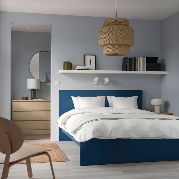 Modern bedroom: blue MALM bed, white linens, wooden dresser, hanging light. Tall headboard, minimalist decor, mirror, chair, side table with lamp.