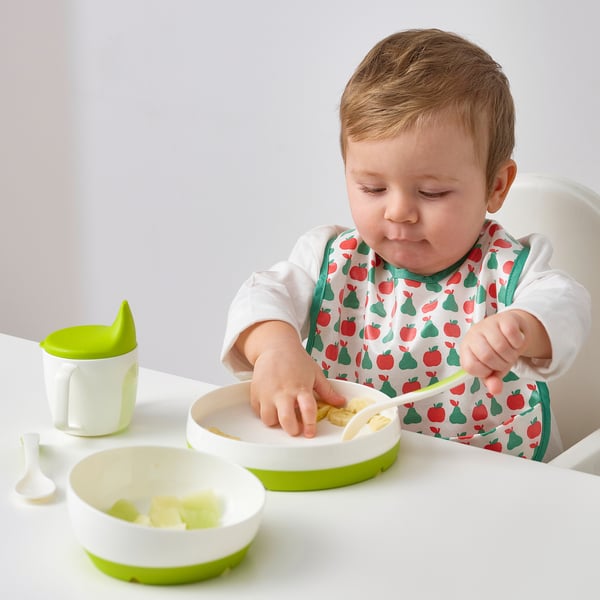 Baby in high chair with fruit bib, eating from bowl with spoon.