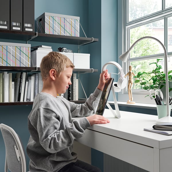 A boy uses a tablet on a desk with a stand holding the tablet. The desk has a lamp, plant, and other items. The wall has shelves with books and boxes.