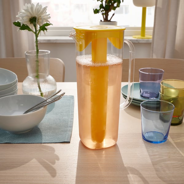 Lemonade table with yellow jug, blue cups, flower vase, sunlight.