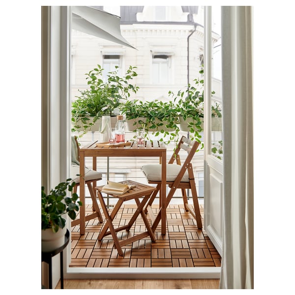 Bright balcony with wooden NÄMMARÖ furniture set. Table and chairs have beige cushions, surrounded by green plants.