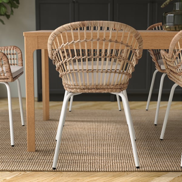 Three hand-woven rattan chairs with white metal legs around a wooden dining table on a woven rug.