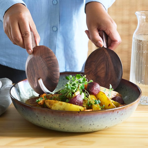 Person holding dark walnut salad servers, serving a colourful vegetable dish in a speckled bowl on a wooden table.