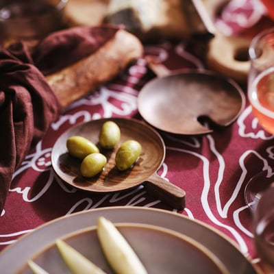 Small wooden dish with olives, part of OLIVTRAST dark walnut serving set, styled on patterned tablecloth.