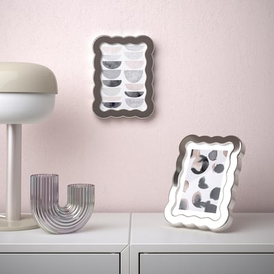 White shelf with silver decorative vases and two framed abstract art pieces. One frame leans on the shelf, the other hangs on a pink wall.
