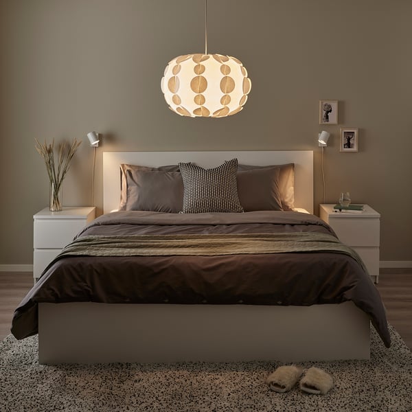 Modern bedroom with a large bed, dark bedding, and a prominent PEKTOLIT pendant lamp with a circular shade, creating a cosy atmosphere.