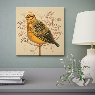 A wall art depicting a yellow bird on a branch, mounted above a table with a lamp, books, and a vase with greenery.