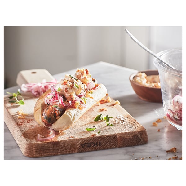 Small wooden cutting board with handle. Holds gourmet hot dog with onions and sauce. Glass with onion slices and spoon. Marble countertop.