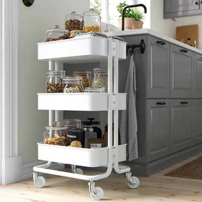 Small white RÅSKOG trolley with three shelves, storing various food jars, near grey kitchen cabinets.