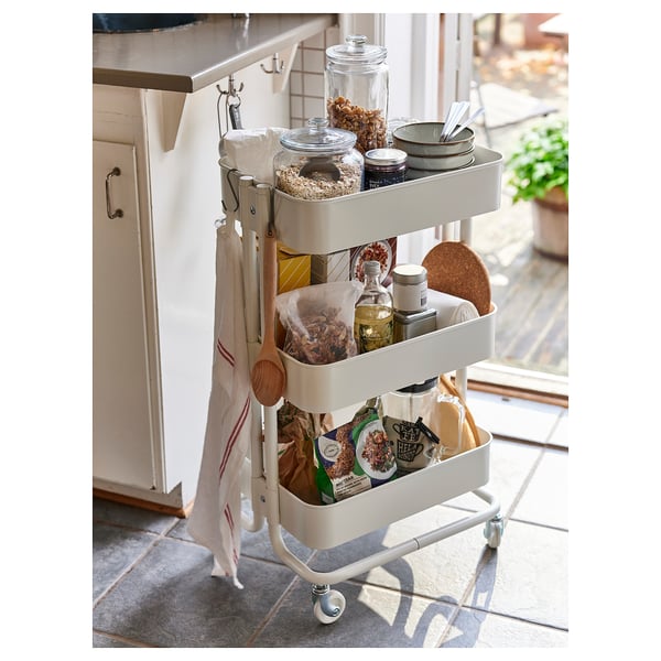 Small white kitchen trolley, RÅSKOG, with three shelves organising food items, kitchen tools, and bags, positioned near a cabinet.