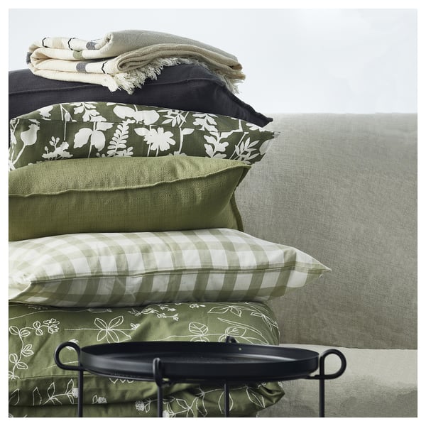 Bed with ROTVECKLARE green, floral pillow. Two green pillows, one checked. Grey blanket, black tray on bed.