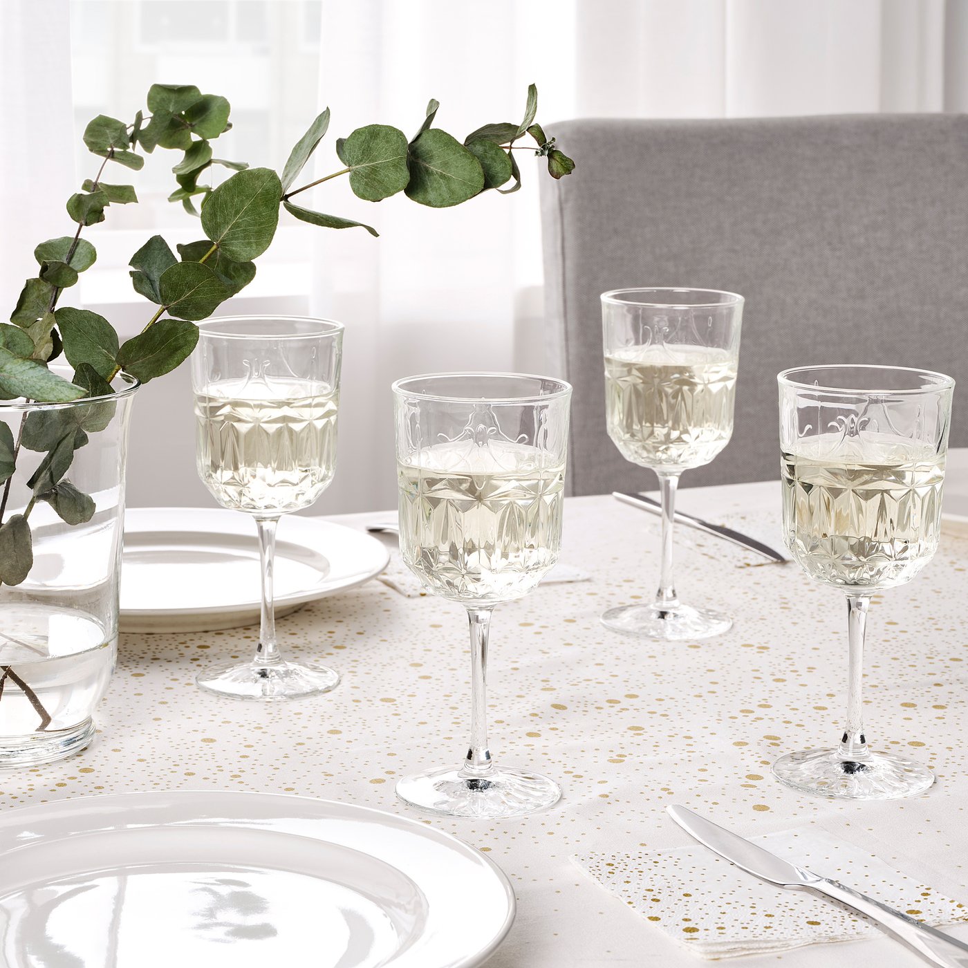 Elegant table setting with ornate glasses, white plates, gold-speckled table runner, and eucalyptus branch.